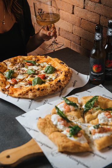 Two pizzas and craft beer on a table with a person holding a wine glass.