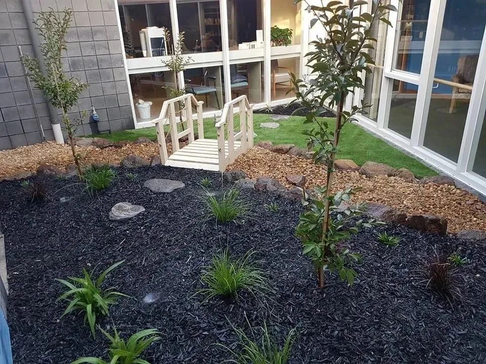 A modern indoor garden with a small wooden bridge and black mulch.