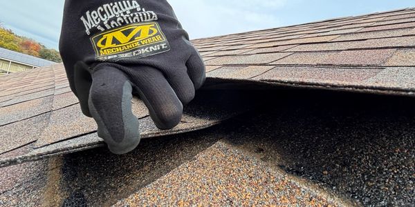 A gloved hand lifting a shingle on a roof, revealing a gap underneath.