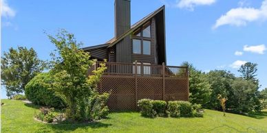 A-frame house with a wooden deck surrounded by green grass and trees under a blue sky.