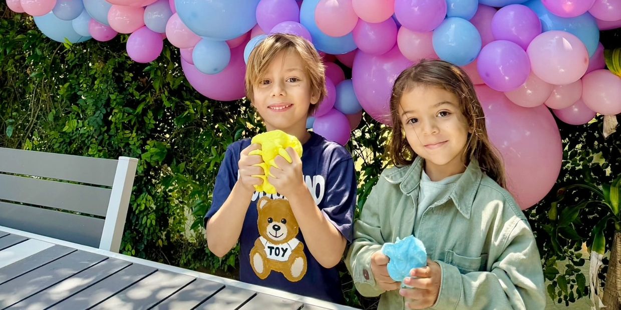 Two children playing with colorful slime under pastel balloons outdoors.