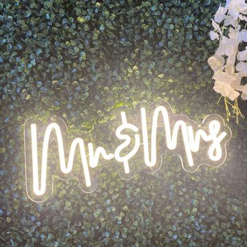 Glowing "Mr & Mrs" sign against a lush green foliage backdrop with white flowers.