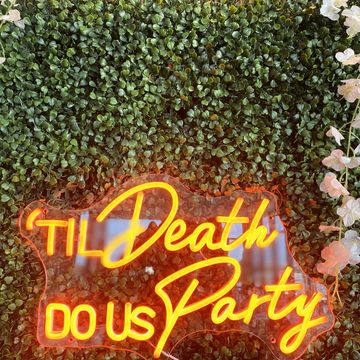 Neon sign reading 'TIL Death DO US Party' against green foliage with white flowers.