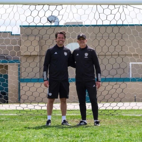 Two men in sportswear standing in front of a soccer goal, smiling.