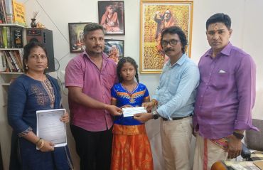 Five people in a room, a girl receiving a document or cheque from two men.