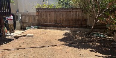 A dry backyard with a wooden fence and two people working near the house.