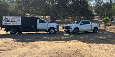 Two white demolition trucks parked on a dirt lot under trees.