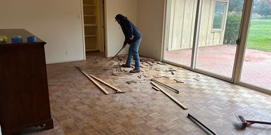 A person removing old parquet flooring in a room with large sliding glass doors.