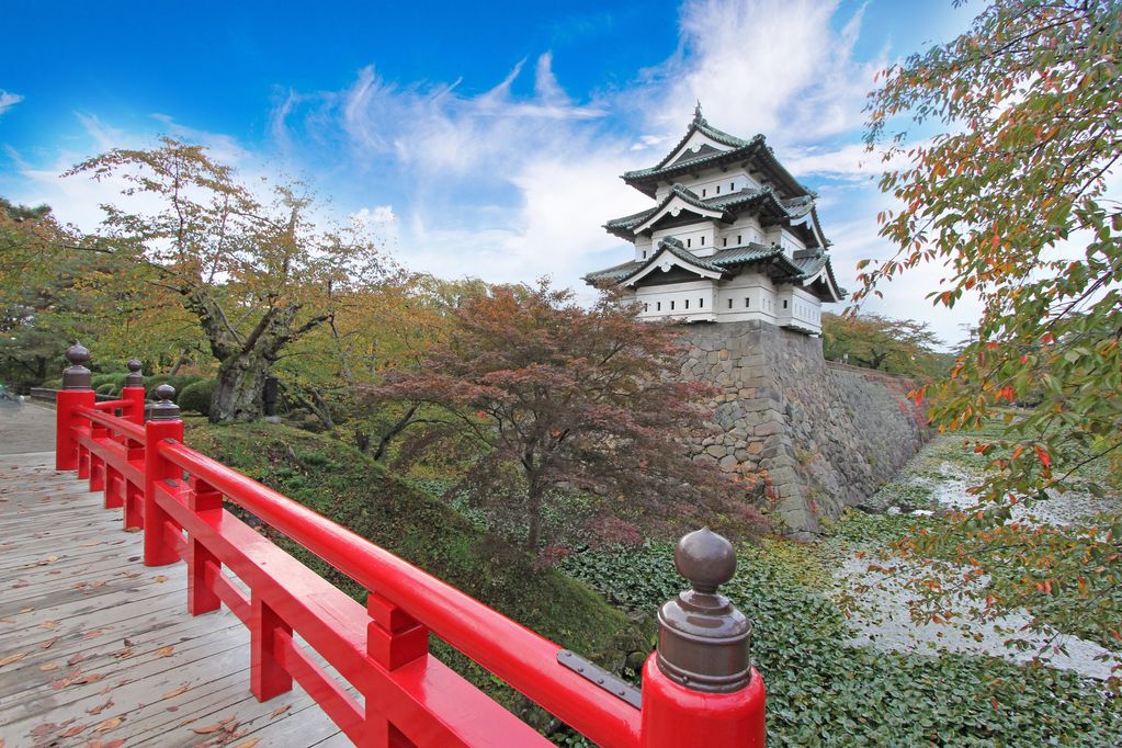A traditional Japanese castle with a bright red bridge in the foreground under a blue sky.