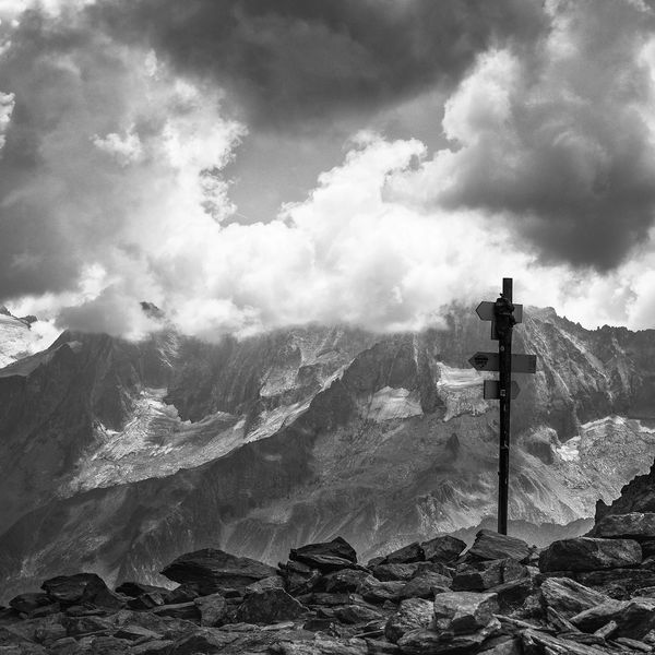 A black  & white image of stony mountains topped by clouds. A signpost has pointers to left & right