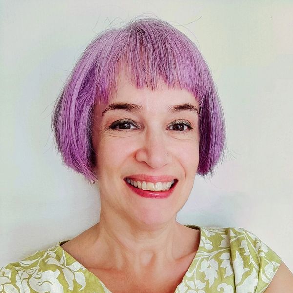 Claire is smiling at the camera. She is a White woman with purple bobbed hair, wearing a green dress