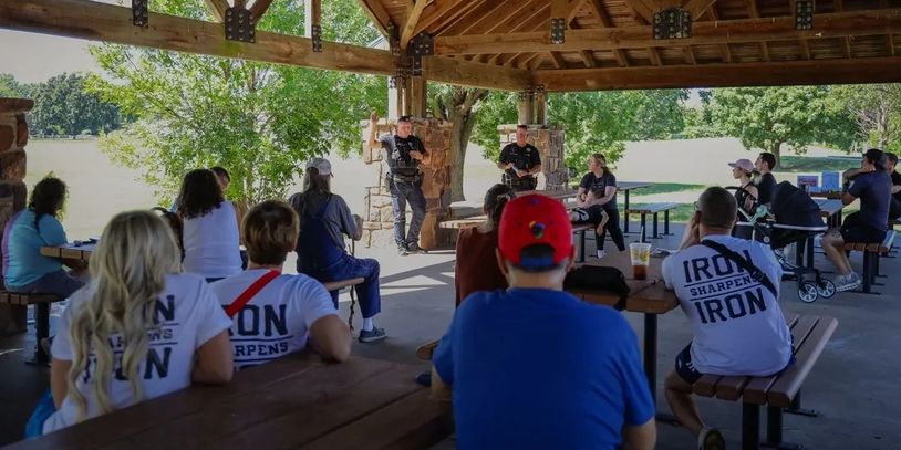 People listen to police officers speaking under a pavilion in a park.