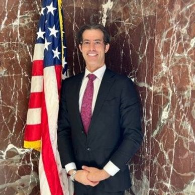 Man in a suit standing beside an American flag with a marble wall background.