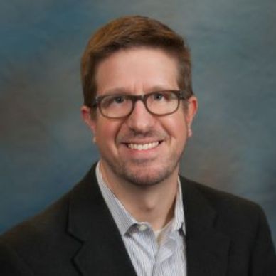 Professional portrait of a smiling man wearing glasses and a suit jacket.