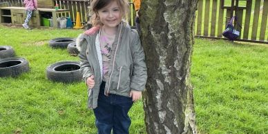 A young girl standing next to a tree in a grassy playground.