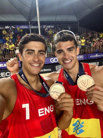 Two male athletes in red ENG jerseys proudly show gold medals at a volleyball event.
