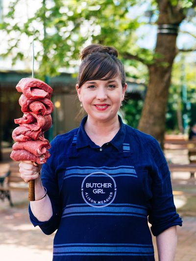 Butcher Girl Alison stands in her apron whilst holding a large skewer of raw beef