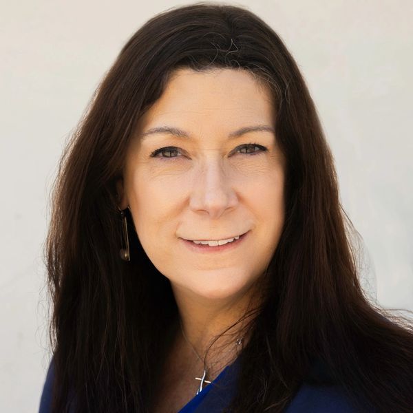 Confident woman with long dark hair wearing a blue blazer and cross necklace.