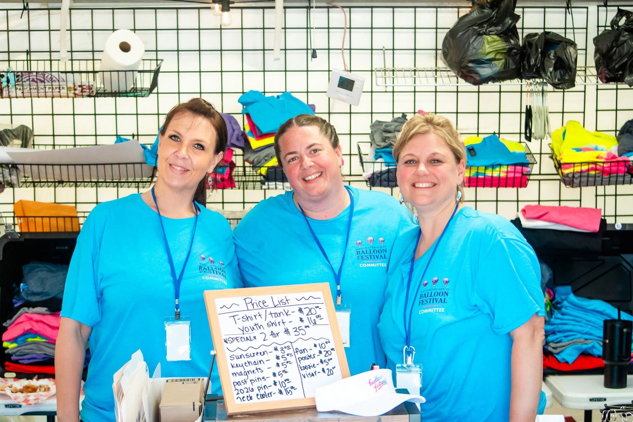 Three women in blue shirts smiling behind a price list at a balloon festival booth.