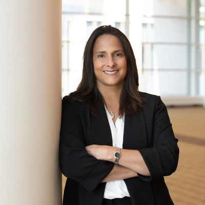 A Latina smiling while wearing a white blouse and black blazer while leaning on a pillar with her ar