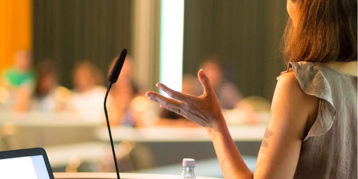 Woman with brown shoulder length hair standing at podium speaking to a group 