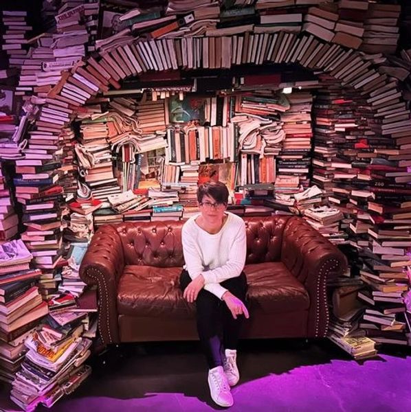 Person sitting on a brown leather couch surrounded by stacks of books.