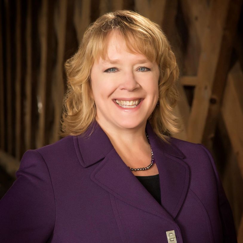 Smiling woman in purple blazer with necklace outdoors.