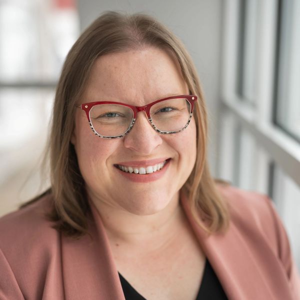Smiling woman with red glasses and a pink blazer.