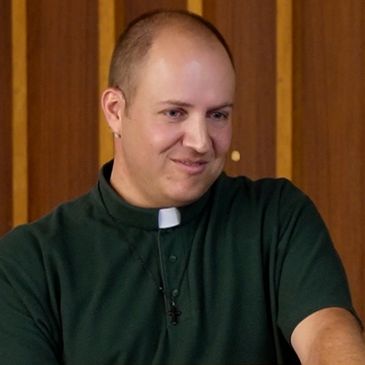 A smiling man wearing a clerical collar and green shirt.
