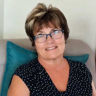Smiling woman with glasses sitting on a chair with blue pillows.