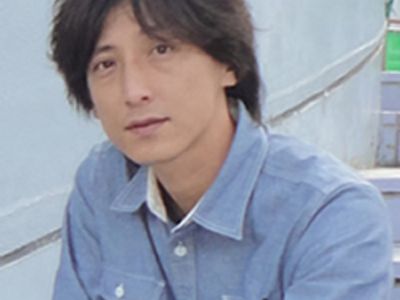 A man with dark hair wearing a blue shirt, sitting outdoors.