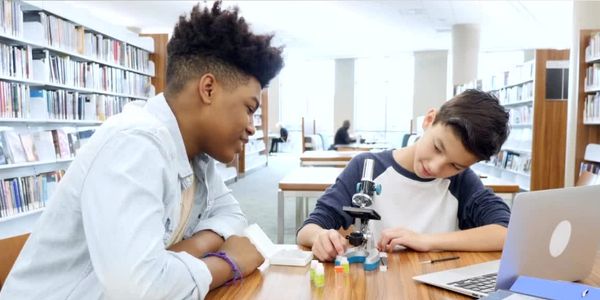 Two students working together on a science project with a microscope in a library.