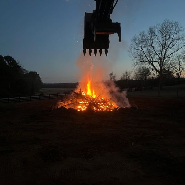 Excavator stoking a fire with trees that were cleared.