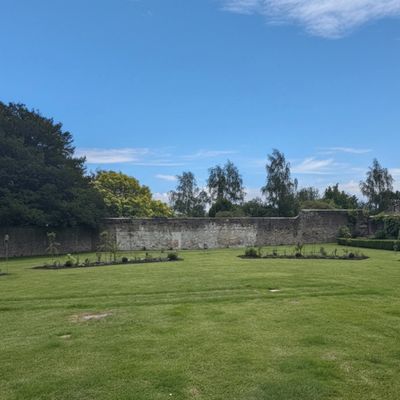 A peaceful garden with manicured grass, flower beds, and a stone wall under a blue sky.