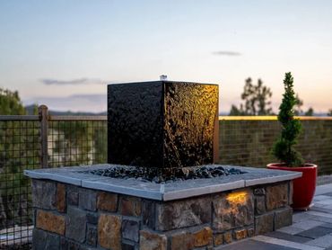 Modern black cube water fountain on a stone base at sunset.