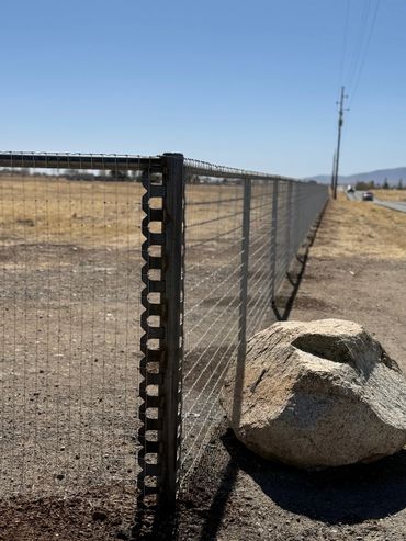 A metal wire fence stretches alongside a road with a large rock resting near it.