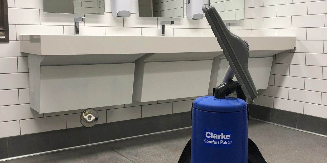 Blue backpack vacuum cleaner in a clean, tiled restroom with sinks, mirrors, and soap dispensers.