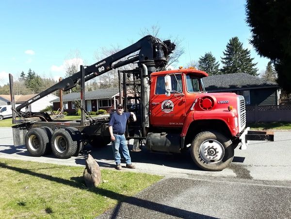 Man standing next to a large red logging truck with a crane on a suburban street.