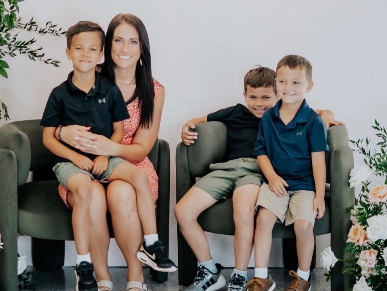 A smiling woman with three boys sitting on chairs surrounded by flowers.