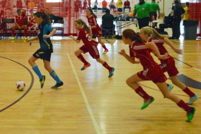 Girls' indoor soccer match with players sprinting towards the ball.