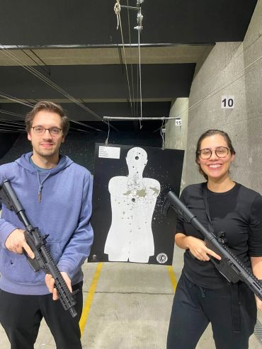 Two people holding rifles at an indoor shooting range with a bullet-riddled target.
