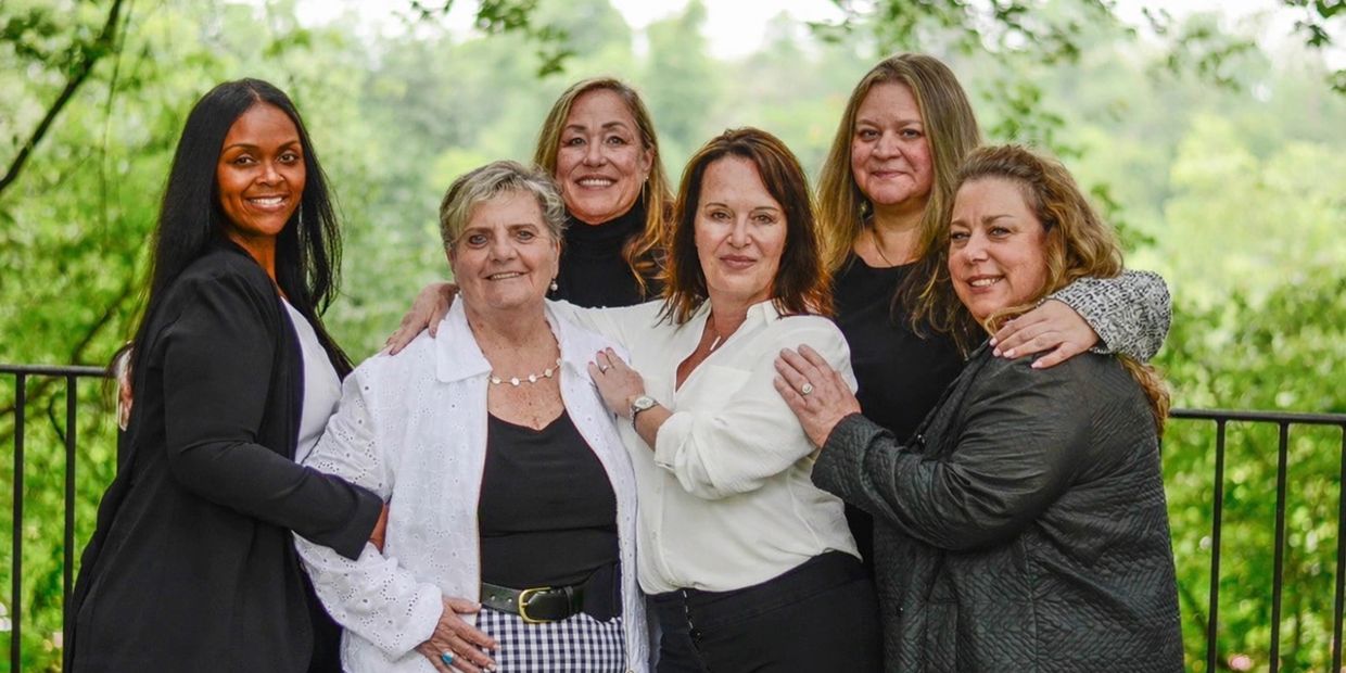 Group of six women smiling outdoors with greenery in the background.