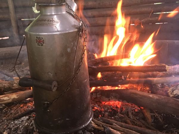 Metal kettle heating over an open wood fire inside a rustic shelter.