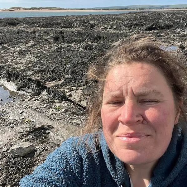 A woman in a blue fleece jacket takes a selfie at a rocky beach on a sunny day.