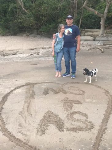 Couple and dog standing by a heart drawn in sand with initials.