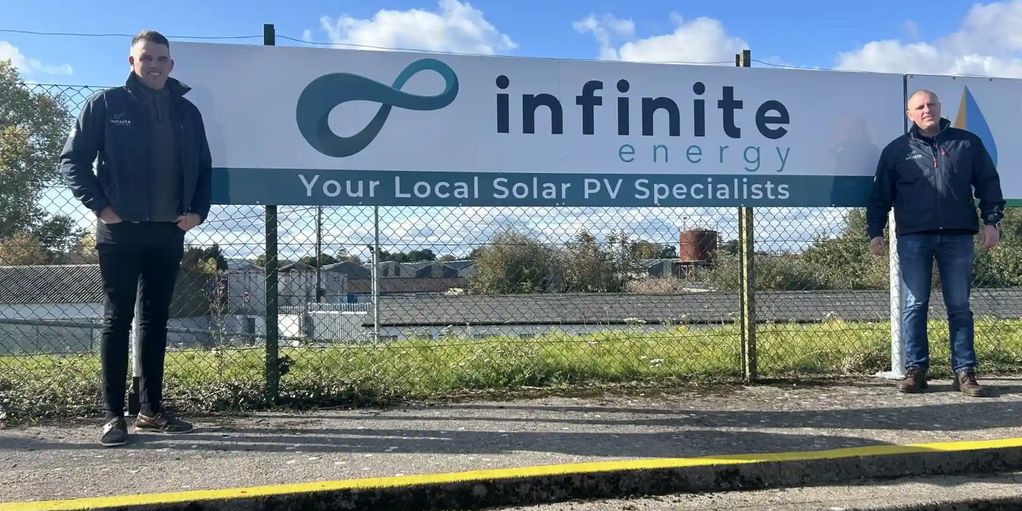 Two men stand beside an Infinite Energy sign outdoors.