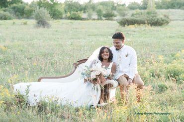 Bride and groom sitting on a vintage couch in a field, smiling.