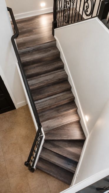 Top-down view of a wooden staircase with black iron railings and tiled flooring below.
