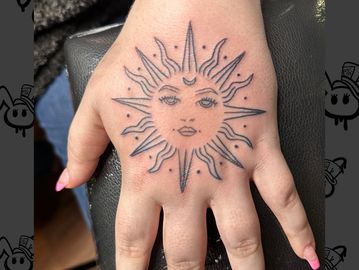 Black ink sun tattoo with a face on the back of a hand.