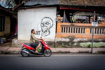 Man riding a red scooter past a wall with skull graffiti.